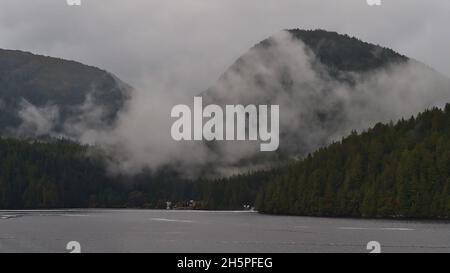 Wilderness landscape with abandoned buildings and clouds rising from dark hills covered by dense forests at the Inside Passage. Stock Photo