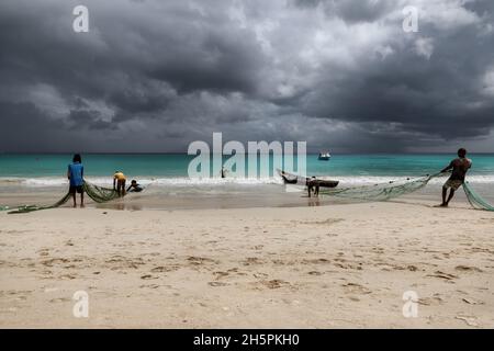 Fishermen pull nets from the sea on a beach in the tropics with dramatic sky on Mahe Island, Seychelles Stock Photo