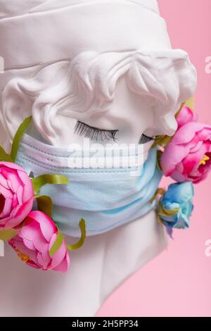 Sculpture of antique girl made of plaster in medical mask with flowers against pink background coronavirus pandemic COVID. Stock Photo