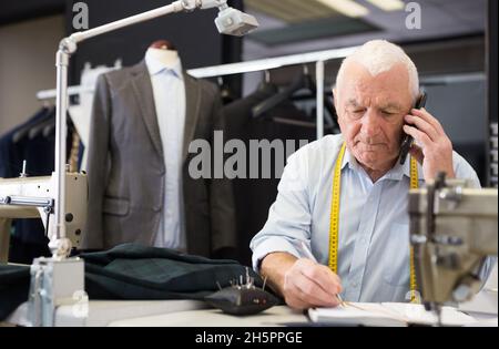 Mature tailor wearing protective mask talking on a mobile phone and taking notes in notebook Stock Photo