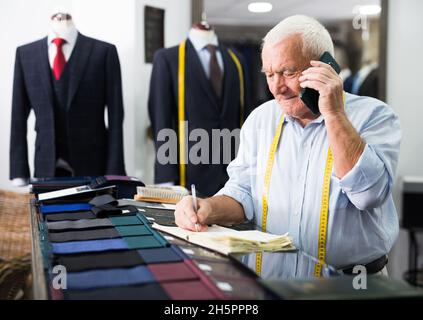 Professional tailor communicates with the client on mobile phone and takes notes in notebook Stock Photo