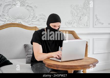 portrait of unrecognizable young hacker in black mask using laptop. Concept of cyber security and data protection. Stock Photo
