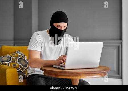 portrait of unrecognizable young hacker in black mask using laptop. Concept of cyber security and data protection. Stock Photo