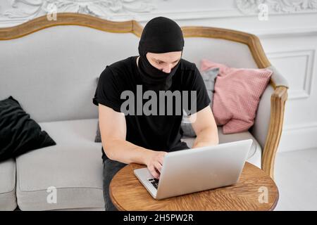 portrait of unrecognizable young hacker in black mask using laptop. Concept of cyber security and data protection. Stock Photo