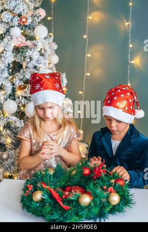 Two children boy and girl in santa hats sit together in decorated room with Christmas tree. Children make christmas wreath Stock Photo