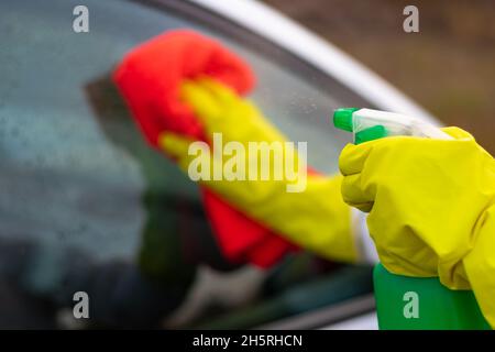 Hands in yellow rubber gloves wipe the glass of a car with a red rag and spray water from a spray gun on a warm autumn day. Wet cleaning. Selective fo Stock Photo