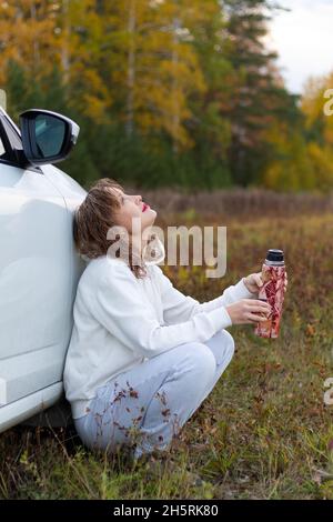 A pretty young woman in a white hoodie sits near a white car with a thermos in her hands and drinks hot tea on a warm autumn day against a background Stock Photo