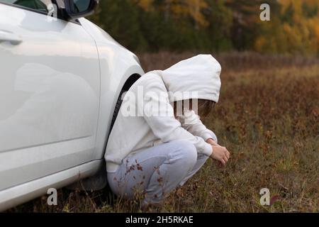 A pretty young woman in a white hoodie and sweatpants sits near a white car with reflection on a warm autumn day against a background of yellow foliag Stock Photo