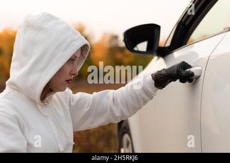 A beautiful young woman in a white sweater with a hood and black gloves opens the car door to steal it. Selective focus. Close-up. Portrait Stock Photo