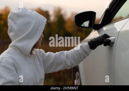 A beautiful young woman in a white sweater with a hood and black gloves opens the car door to steal it. Selective focus. Close-up. Portrait Stock Photo