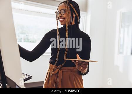Creative painter working on a painting in her art studio. Happy female painter smiling cheerfully while holding a palette and drawing on a canvas. Art Stock Photo