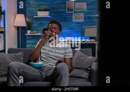 African american young man watching television laughing during comedy movie relaxing in living room. Black guy sitting on sofa while eating popcorn enjoying spending tine alone Stock Photo