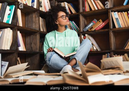 smart african american woman in eyeglasses sitting surrounded by books in library Stock Photo