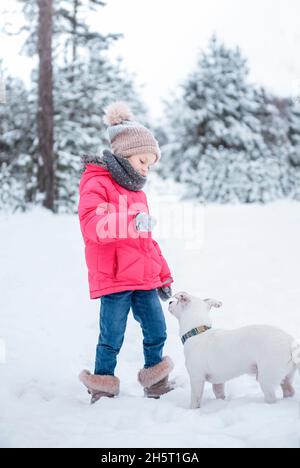 Little girl in a bright jacket plays in the winter snowy forest with her dog jack russell terrier Stock Photo