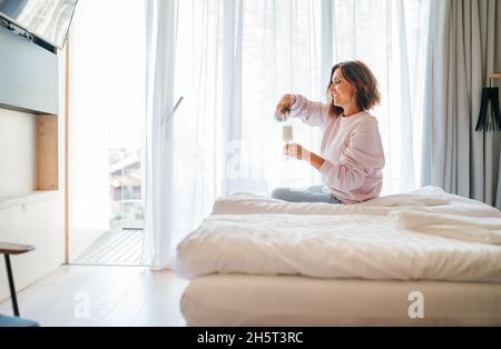 Laughing woman sitting on hotel room bed next to the window and pouring white wine champagne into a tall glass.Event celebration or alcoholism problem Stock Photo