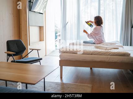 Lonely Woman sitting on a modern furniture hotel room bed next to the window and pouring white wine champagne into a tall glass.Event celebration or a Stock Photo