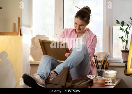 Caucasian woman artist working on a painting in bright daylight studio. Happy artist draws an art project with paints and a brush in the workshop Stock Photo