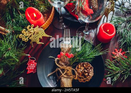 Festive Christmas dinner table setting for two decorated with fir branches, red candles, cones, ornaments. Plates with cutlery and glasses surrounded Stock Photo