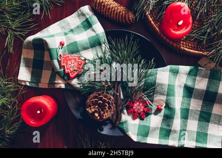 Festive Christmas dinner table setting decorated with fir branches, red candles, cones, ornaments. Black plate with green checkered napkin Stock Photo