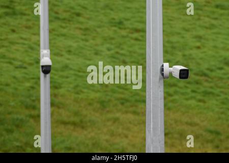 Two white security cameras mounted on a grey pole, covered in grass in the background Stock Photo