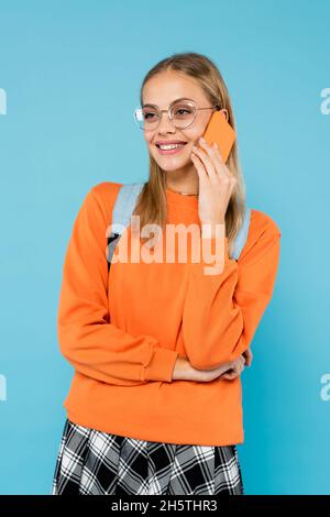 Pretty student in eyeglasses with backpack talking on mobile phone isolated on blue Stock Photo