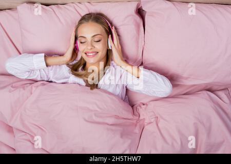 Top view of cheerful woman listening music in headphones while lying on bed Stock Photo