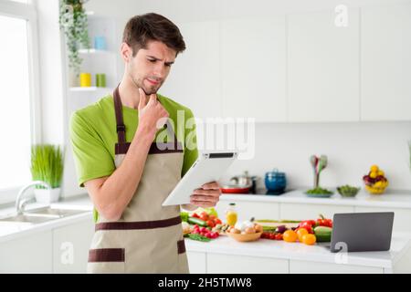 Portrait of attractive minded guy cooking dinner using device watching web video tutorial at home kitchen indoors Stock Photo