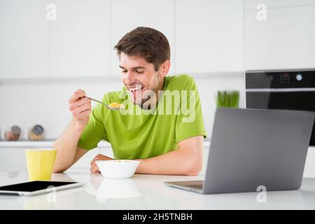 Portrait of attractive cheerful guy eating healthy lunch domestic cuisine watching tv show at light white kitchen home indoors Stock Photo