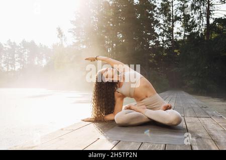A woman practicing yoga, sitting in the lotus position and doing side bends, works out in the park on a sunny morning on a wooden bridge by the pond Stock Photo
