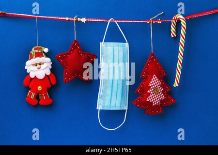Christmas felt decorations and disposable medical mask hang on decorative ribbon on blue background, holiday quarantine concept Stock Photo