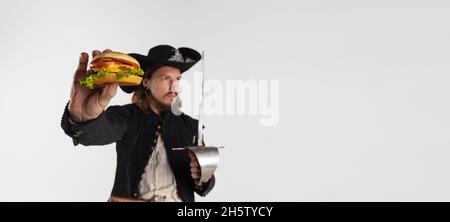 Cropped portrait of brutal man, pirate in vintage costume with sword holding burger isolated over white background. Flyer Stock Photo