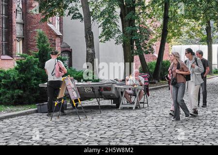 Mature man,glassblower, sitting at table in park, in process of artistic glass processing.Tourists passing by are interesting in glass miniatures Stock Photo