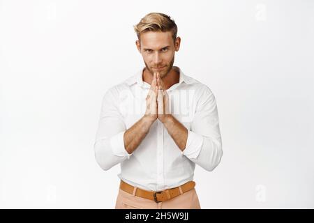 Young devious blond man in fancy clothes, having smth interesting in mind, looking cunning, thinking, scheming something, standing over white Stock Photo