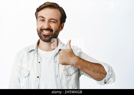 Cheerful caucasian male model with long beard, winking and showing thumbs up, smiling pleased, giving approval, white background Stock Photo