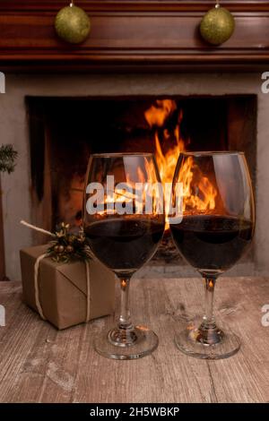 two crystal glasses with red wine next to a box with gift paper and decorated for Christmas in the background a campfire, objects for a romantic date Stock Photo