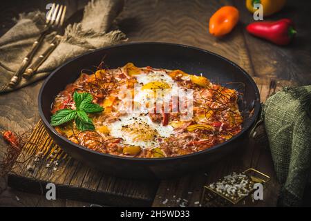 Shakshuka in an iron pan on wooden background. Middle eastern traditional dish. Fried eggs with tomatoes, bell pepper, vegetables and herbs. Stock Photo