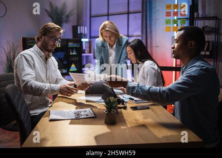 Group of competent multiracial economists gathering late at office room for discussing financial situation of company. Cooperation for common succes. Modern gadgets for work. Stock Photo