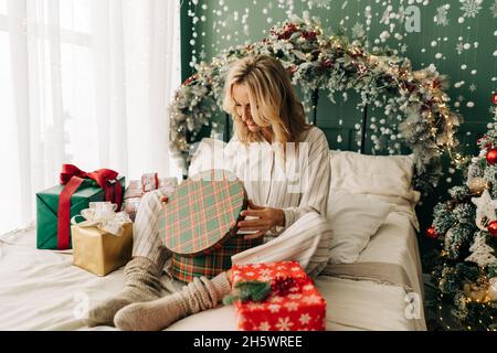 Caucasian happy attractive blonde woman opens Christmas gift boxes while sitting on bed in bedroom. Stock Photo