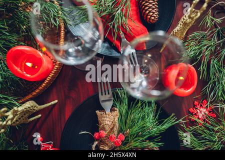Festive Christmas dinner table setting for two decorated with fir branches, red candles, cones, ornaments. Plates with cutlery and glasses surrounded Stock Photo