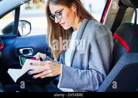 Busy female entrepreneur sitting on passenger seat in car and taking notes in notepad while using smartphone Stock Photo