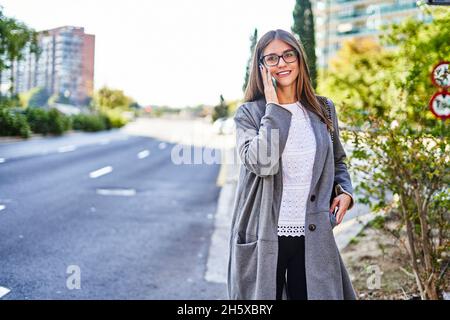 Serious female in formal clothes and coat with handbag crossing asphalt road while talking on cellphone Stock Photo