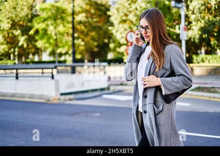 Serious female in formal clothes and coat with handbag crossing asphalt road while talking on cellphone Stock Photo