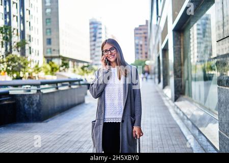 Positive female entrepreneur wearing formal clothes while talking on cellphone and looking away while toothy smile Stock Photo