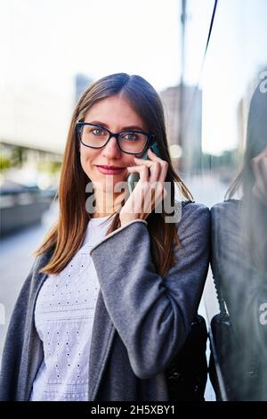 Positive female entrepreneur wearing formal clothes leaning on wall and talking on cellphone and looking at camera Stock Photo