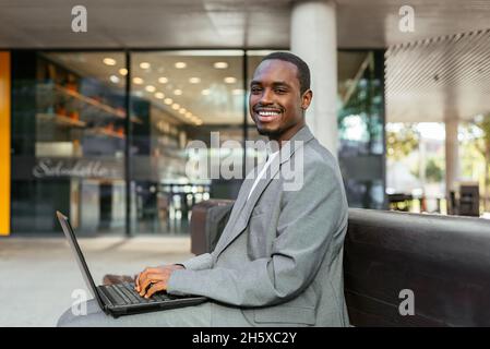 Side view of happy African American male entrepreneur sitting on bench and typing on netbook while working online Stock Photo
