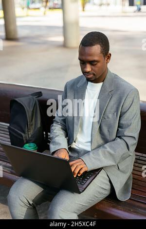 From above of concentrated African American male entrepreneur sitting on bench and typing on netbook while working online Stock Photo