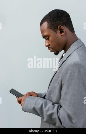 Serious African American businessman in formal wear and earbuds browsing mobile phone and looking away near grey wall Stock Photo