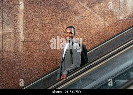 Side view of African American male entrepreneur with backpack listening to songs in TWS earbuds on escalator Stock Photo