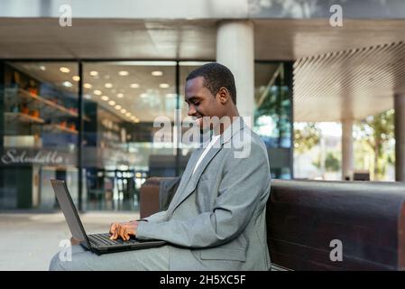 Side view of happy African American male entrepreneur sitting on bench and typing on netbook while working online Stock Photo