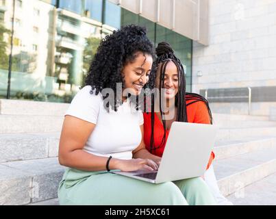Cheerful diverse female friends in trendy clothes sitting on stair and browsing laptop on city street in summer day Stock Photo